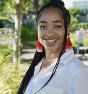 Mysia Pierce Lewis smiling for her headshot with greenery in the background and red earrings.