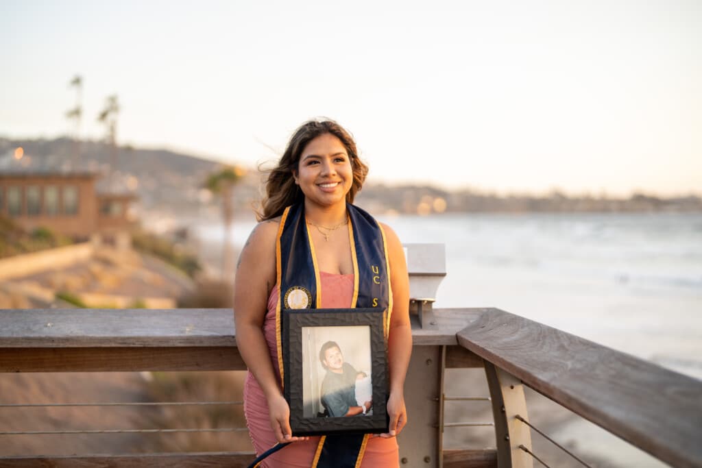 Photo Allison with beach in background in UC San Diego graduation stole holding framed photo of father 