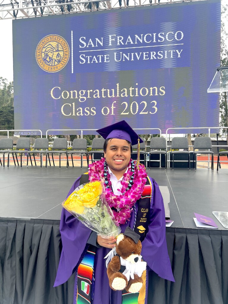 Alex Aceves with his cap and gown holding flowers at his SFSU graduation