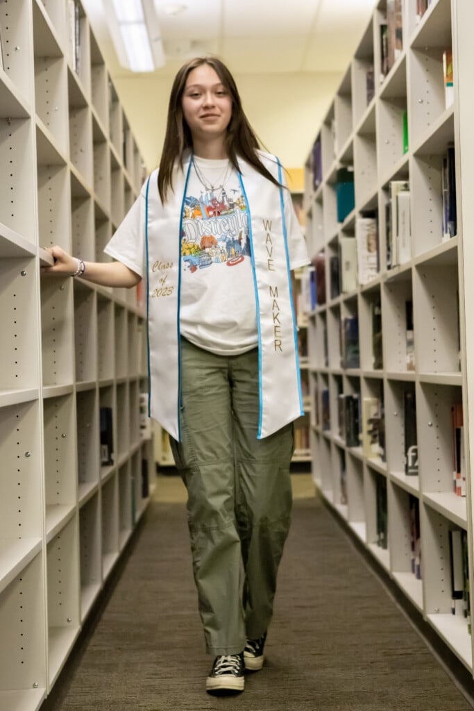 Dayan Guzman stands in between two library stacks and poses with a White Making Waves stole