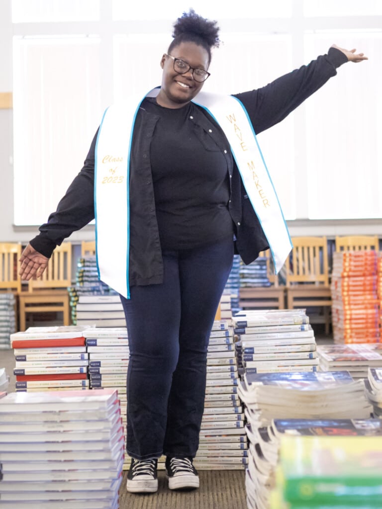 Jazaria DeVoe-Harris stands in between stacks of books in a library and poses with a white Making Waves stole