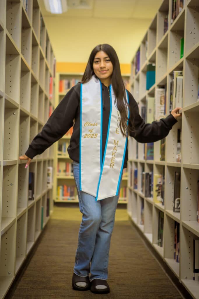 Isabella-Lopez stands in between two library stacks and psoes with a white Making Waves stole