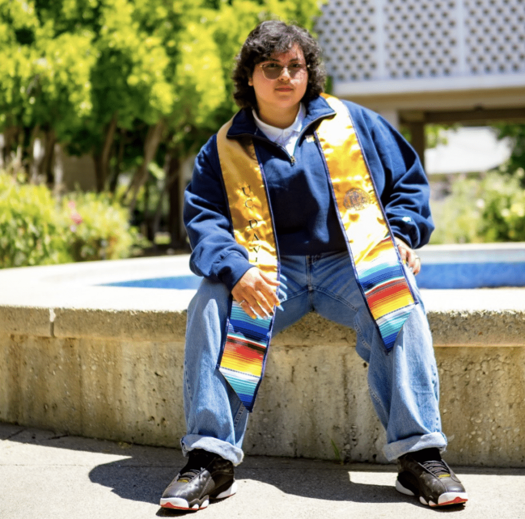 Alejandro Diaz sitting at fountain leaning forward wearing colorful graduation stole 