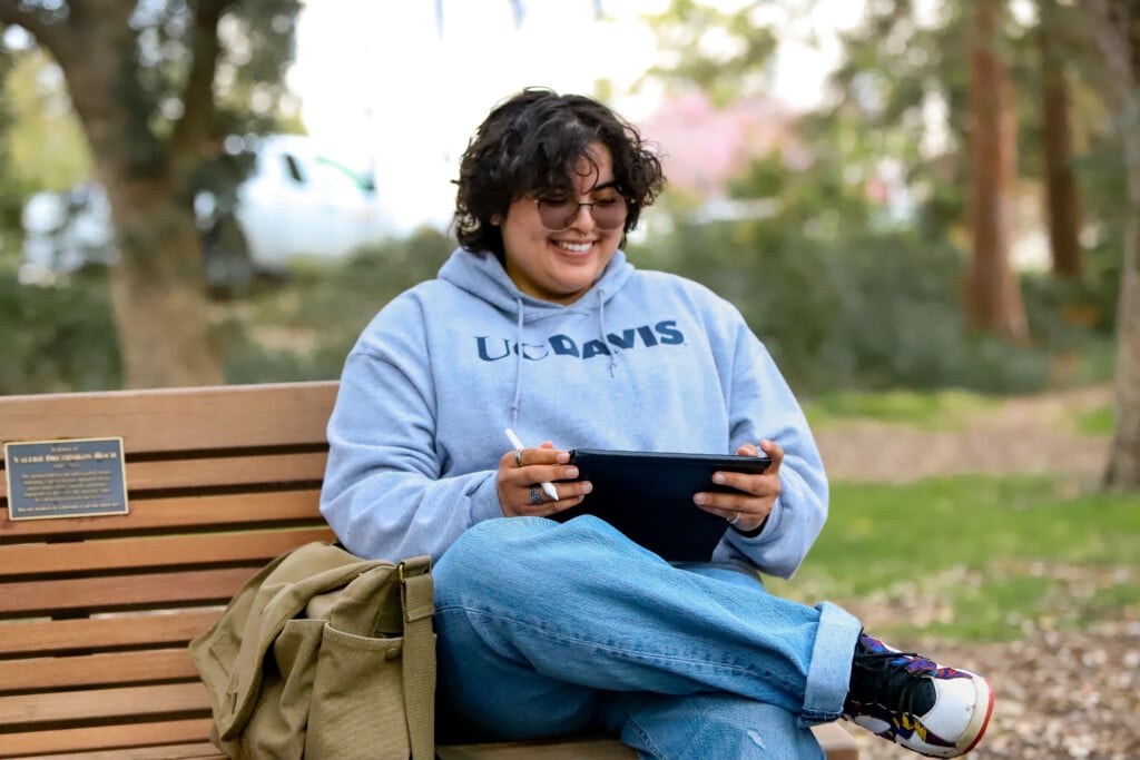 Alejandro wearing UC Davis sweater sitting on bench with tablet smiling