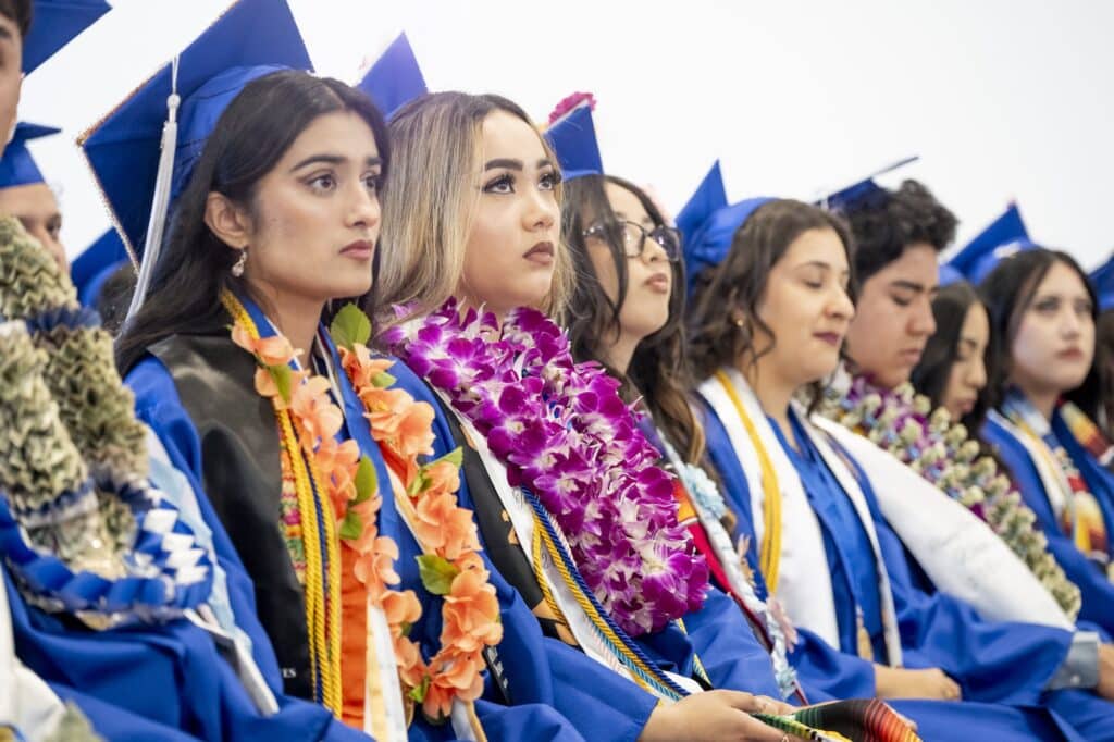 Students wearing blue caps and gowns and flower leis listening to speakers