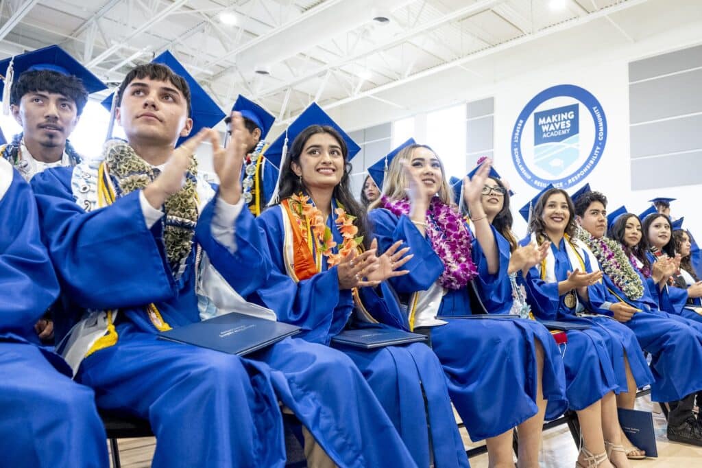 Students cheering in blue caps and gowns in seats at graduation event