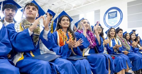 Students cheering in blue caps and gowns in seats at graduation event